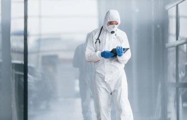 Laboratuvar önlüklü, koruyucu gözlüklü ve maskeli bir erkek doktor elinde not defteri tutuyor..