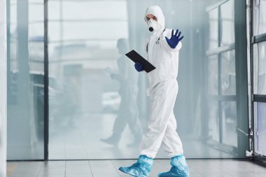 Shows stop gesture by hand. Male doctor scientist in lab coat, defensive eyewear and mask holding notepad in hands.
