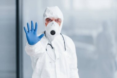 Shows stop gesture by hand. Male doctor scientist in lab coat, defensive eyewear and mask.