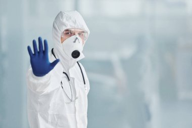 Shows stop gesture by hand. Male doctor scientist in lab coat, defensive eyewear and mask.