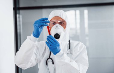 Laboratuvar önlüklü, koruyucu gözlüklü ve maskeli erkek doktor test tüpünü kanla tutuyor..