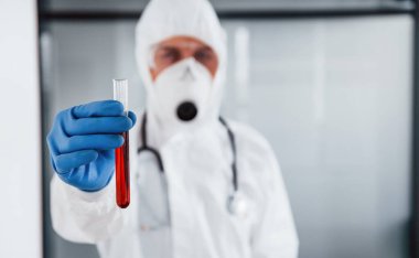Laboratuvar önlüklü, koruyucu gözlüklü ve maskeli erkek doktor test tüpünü kanla tutuyor..