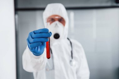 Laboratuvar önlüklü, koruyucu gözlüklü ve maskeli erkek doktor test tüpünü kanla tutuyor..