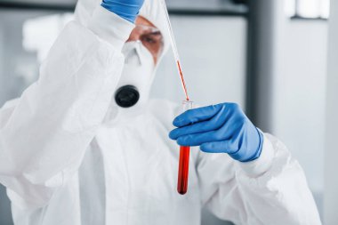 Laboratuvar önlüklü, koruyucu gözlüklü ve maskeli bir erkek doktor, içinde kan olan test tüpüyle çalışır..