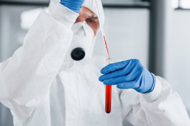 Laboratuvar önlüklü, koruyucu gözlüklü ve maskeli bir erkek doktor, içinde kan olan test tüpüyle çalışır..