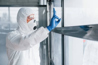 Laboratuvar önlüklü, koruyucu gözlüklü ve maskeli bir erkek doktor pencereden içeri girerken....