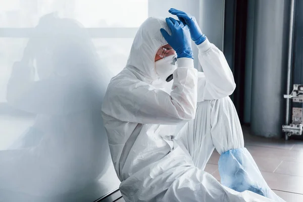 身穿实验室外套 戴防卫性眼镜和面具的疲惫的医生科学家坐着休息 — 图库照片