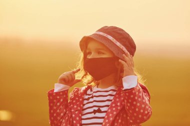 Dışarıda turuncu güneş ışığıyla aydınlatılan siyah koruyucu maskeli şirin küçük kızın portresi..