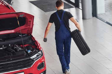 Siyah-mavi üniformalı işçi araba direksiyonu tutuyor ve kırmızı otomobilin yanında yürüyor..