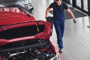 Siyah-mavi üniformalı işçi araba direksiyonu tutuyor ve kırmızı otomobilin yanında yürüyor..