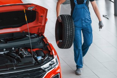 Siyah-mavi üniformalı işçi araba direksiyonu tutuyor ve kırmızı otomobilin yanında yürüyor..