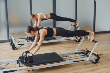 With help of gym equipment. Two women in sportive wear and with slim bodies have fitness yoga day indoors together.
