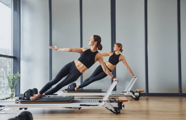 Spor aletlerinin üzerinde durup esneme hareketleri yapmak. Spor elbiseli ve zayıf vücutlu iki kadın kapalı alanda spor yoga yapıyorlar..