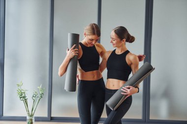 Ellerinde paspaslarla dikiliyorlar. Spor elbiseli ve zayıf vücutlu iki kadın kapalı alanda spor yoga yapıyorlar..