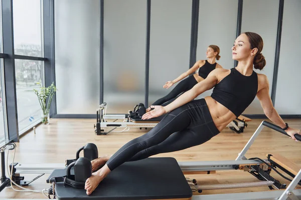 Spor aletlerinin üzerinde durup esneme hareketleri yapmak. Spor elbiseli ve zayıf vücutlu iki kadın kapalı alanda spor yoga yapıyorlar..