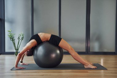 Fitness topunu kullanarak. Spor elbiseli ve zayıf vücutlu genç bir kadın evde yoga yapıyor..