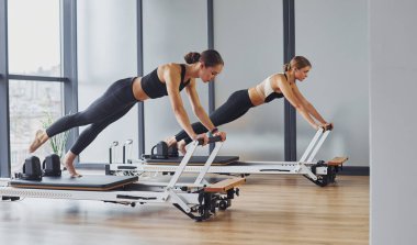 Spor aletlerini kullanarak. Spor elbiseli ve zayıf vücutlu iki kadın kapalı alanda spor yoga yapıyorlar..