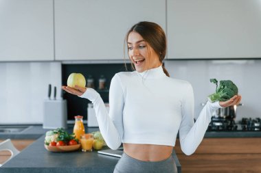 Poses for a camera with vegetables. Young european woman is indoors at kitchen indoors with healthy food.
