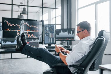 Sandalyeye oturur. Genç borsacı erkek ofis içinde çalışıyor. Ekranların çoğu.