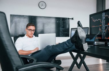 Sandalyeye oturur. Genç borsacı erkek ofis içinde çalışıyor. Ekranların çoğu.