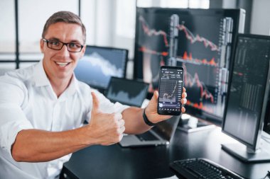 Telefonu tutuyor. Genç borsacı erkek ofis içinde çalışıyor. Ekranların çoğu.
