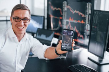Telefonu tutuyor. Genç borsacı erkek ofis içinde çalışıyor. Ekranların çoğu.