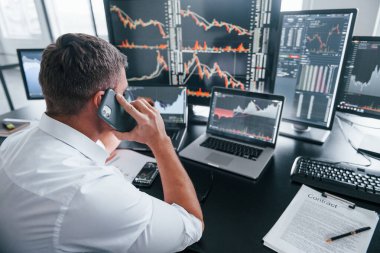 Telefonla konuşuyor. Genç borsacı erkek ofis içinde çalışıyor. Ekranların çoğu.