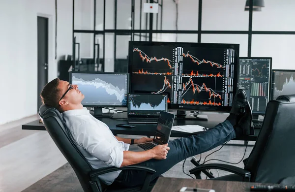 Sandalyeye oturur. Genç borsacı erkek ofis içinde çalışıyor. Ekranların çoğu.