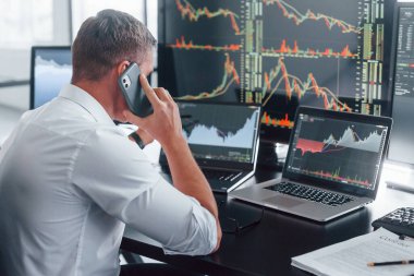 Telefonla konuşuyor. Genç borsacı erkek ofis içinde çalışıyor. Ekranların çoğu.