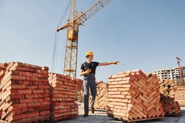 Shows gestures and talks. Construction worker in uniform and safety equipment have job on building.