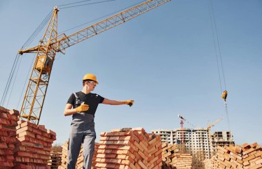 Shows gestures and talks. Construction worker in uniform and safety equipment have job on building.