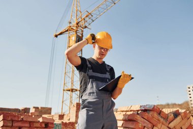Holds notepad with documents. Construction worker in uniform and safety equipment have job on building.