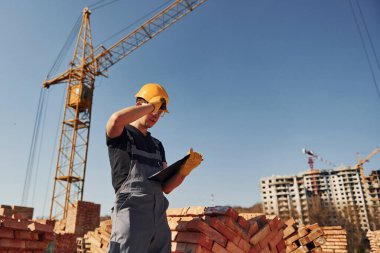 Holds notepad with documents. Construction worker in uniform and safety equipment have job on building.