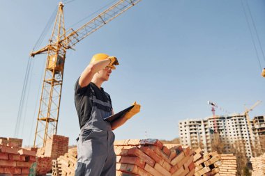 Holds notepad with documents. Construction worker in uniform and safety equipment have job on building.
