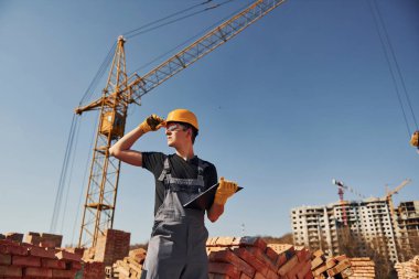 Holds notepad with documents. Construction worker in uniform and safety equipment have job on building.