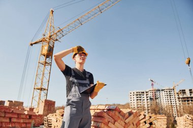 Holds notepad with documents. Construction worker in uniform and safety equipment have job on building.