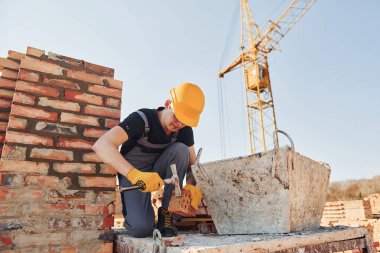 Holding brick and using hammer. Construction worker in uniform and safety equipment have job on building.