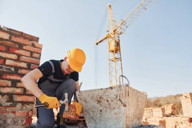 Holding brick and using hammer. Construction worker in uniform and safety equipment have job on building.