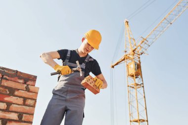 Holding brick and using hammer. Construction worker in uniform and safety equipment have job on building.
