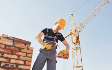 Holding brick and using hammer. Construction worker in uniform and safety equipment have job on building.