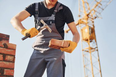 Holding brick and using hammer. Construction worker in uniform and safety equipment have job on building.