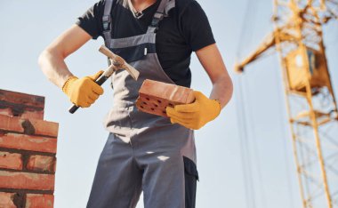Holding brick and using hammer. Construction worker in uniform and safety equipment have job on building.