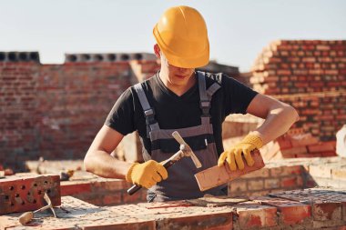 Holding brick and using hammer. Construction worker in uniform and safety equipment have job on building.