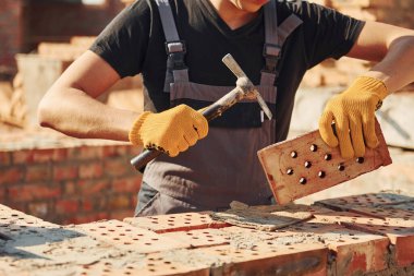 Holding brick and using hammer. Construction worker in uniform and safety equipment have job on building.
