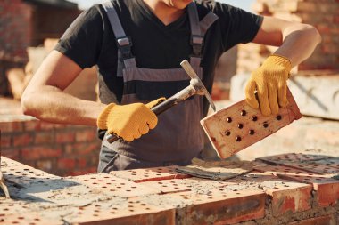 Holding brick and using hammer. Construction worker in uniform and safety equipment have job on building.