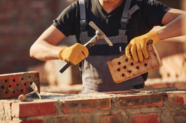 Holding brick and using hammer. Construction worker in uniform and safety equipment have job on building.