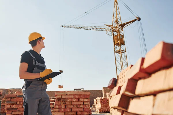 Holds notepad with documents. Construction worker in uniform and safety equipment have job on building.