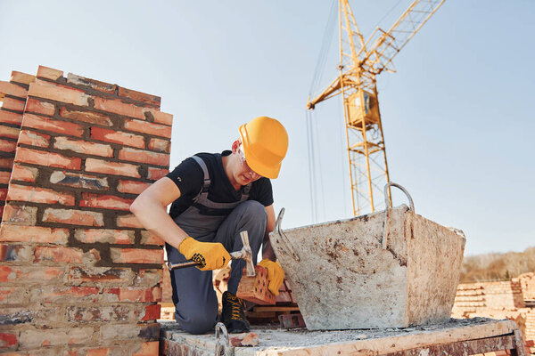 Держать кирпич и молотком. Строитель в форме и технике безопасности имеет работу по строительству.