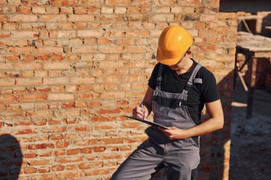 Holds notepad with documents. Construction worker in uniform and safety equipment have job on building.