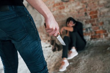 Violent man standing with knife in hand and threatens girl that sits on the floor in abandoned building.